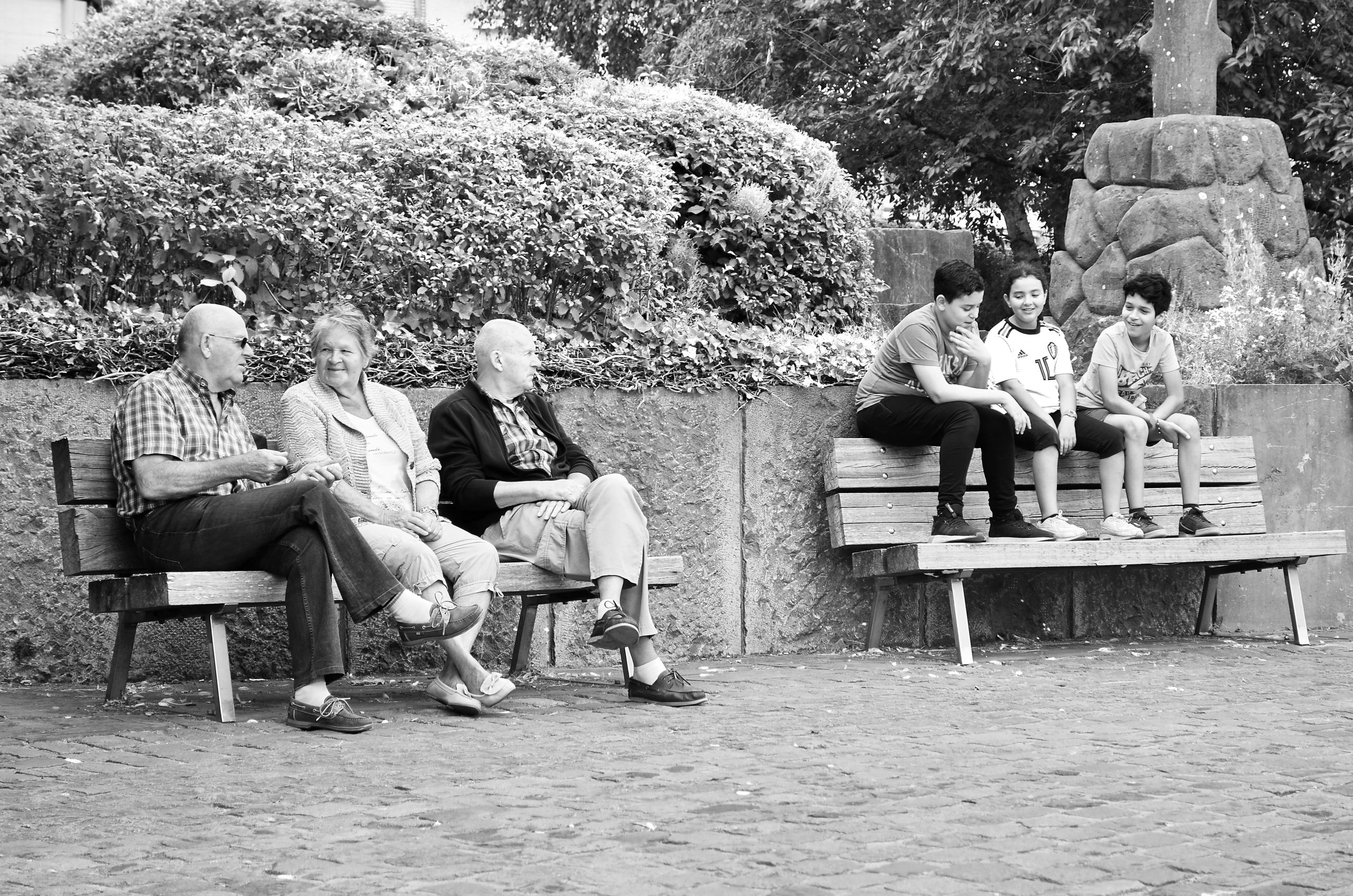 Photo noir et blanc avec 3 personnes agées sur un banc et 3 jeunes sur un autre banc